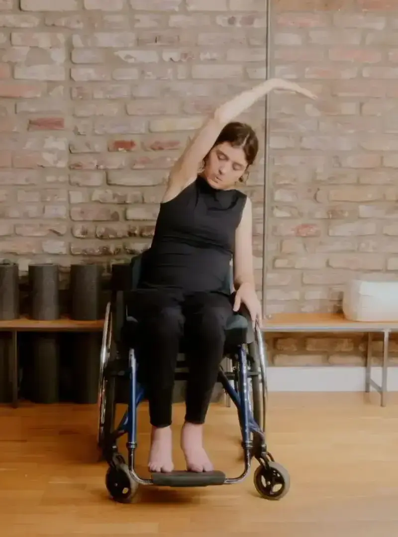 Mechthild sitzt im Rollstuhl und trägt ein schwarzes Top und eine schwarze Leggings. Sie streckt einen Arm über den Kopf. Der andere liegt auf dem Rolli ab.